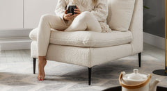 Crèmekleurige bouclé fauteuil Cute met zwarte poten, waarop een persoon in bijpassende kleding zit, genietend van een warme drank. De stoel staat op een lichtgekleurd, geometrisch patroonkleed, en op de voorgrond is een theepot op een dienblad zichtbaar. De setting straalt warmte en gezelligheid uit.