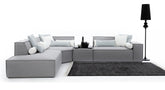 Modulaire, lichtgrijze hoekbank Sense met strakke lijnen en bekleed met kussens in witte en lichtblauwe tinten, waaronder cilindervormige nekkussens. De bank staat op een donkergrijs vloerkleed met lichte randen, en een hoge, zwarte vloerlamp voegt een stijlvol accent toe.