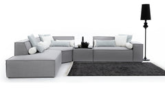 Modulaire, lichtgrijze hoekbank Sense met strakke lijnen en bekleed met kussens in witte en lichtblauwe tinten, waaronder cilindervormige nekkussens. De bank staat op een donkergrijs vloerkleed met lichte randen, en een hoge, zwarte vloerlamp voegt een stijlvol accent toe.