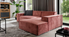 Hoekbank Sense met chaise longue rechts in een warme roze stof, geplaatst in een moderne woonkamer met veel natuurlijk licht. De bank is gecombineerd met een vloerkleed en groene plant, wat de ruimte een eigentijdse en uitnodigende sfeer geeft.