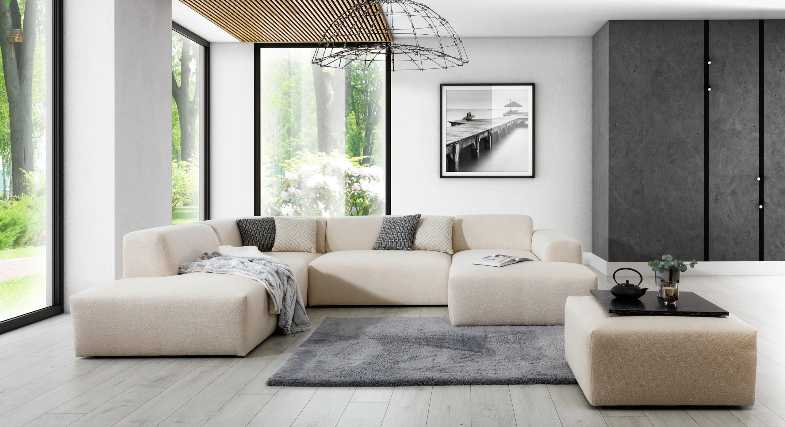 U-bank Vibe met chaise longue rechts in een ruime, moderne woonkamer, gecombineerd met een grijs vloerkleed, zwarte decoratieve tafel en wandkunst. Grote ramen zorgen voor veel natuurlijk licht, waardoor de lichte bekleding van de bank en het minimalistische interieur optimaal tot hun recht komen.
