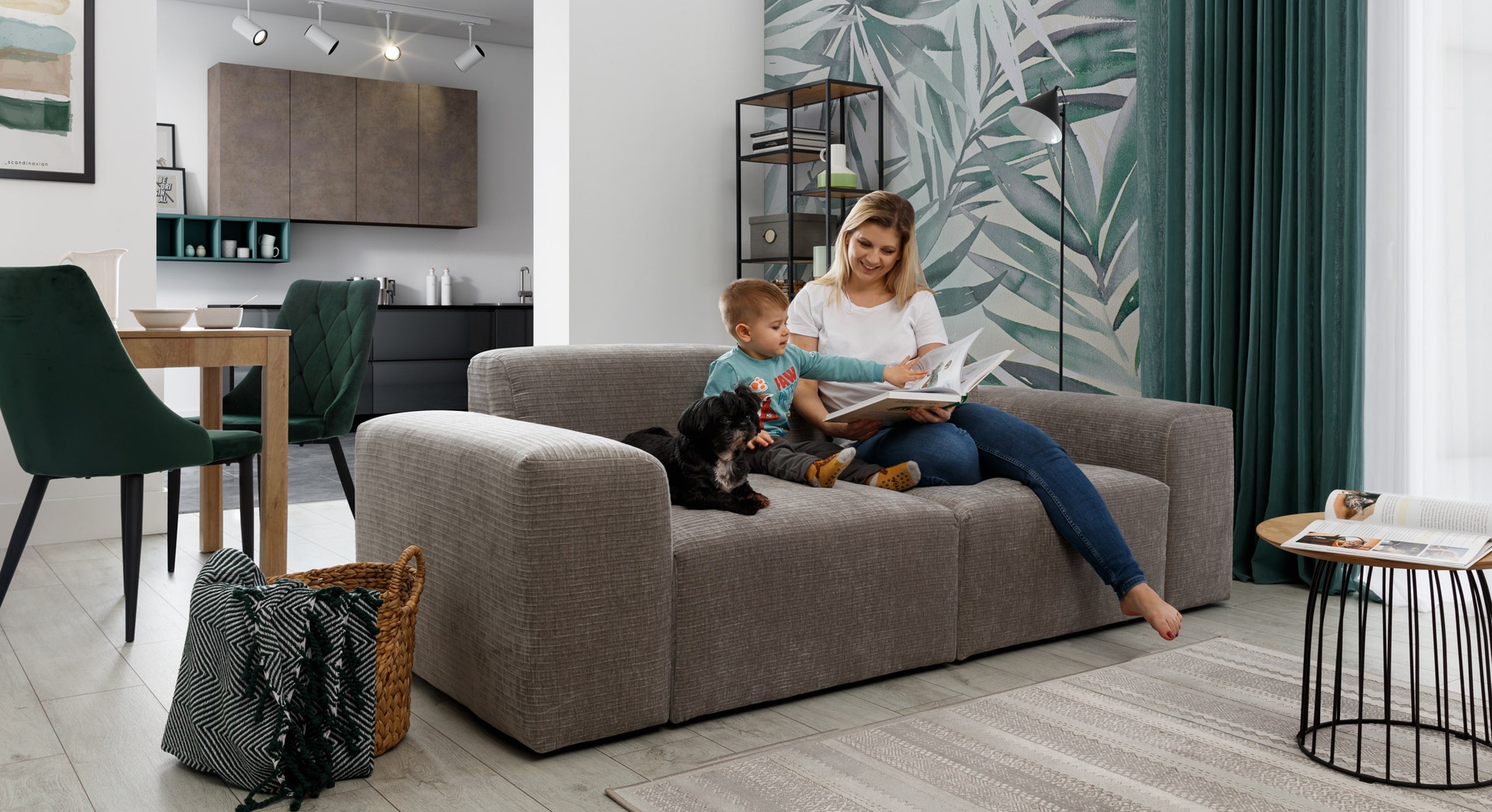 Een moderne woonkamer waarin een vrouw samen met een jong kind op een grijze bank zit en een boek leest, terwijl een kleine zwarte hond naast hen ligt. De achtergrond bevat groene botanische accenten op de muur en gordijnen, een metalen bijzettafel met een tijdschrift, en een mand met een deken naast de bank. De ruimte oogt uitnodigend en gezellig, met een mix van natuurlijke en moderne elementen.