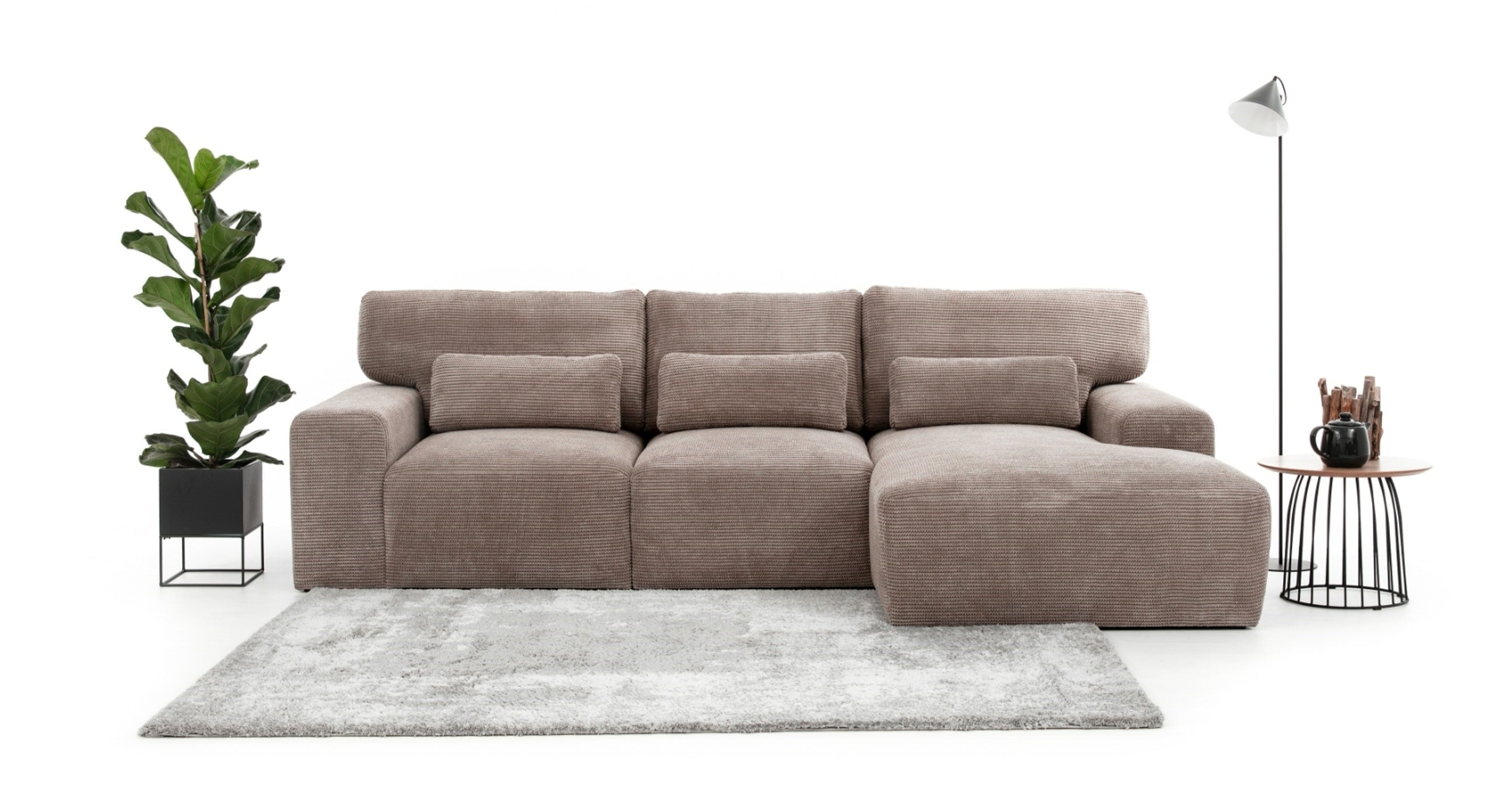 Hoekbank Fun met chaise longue rechts, afgebeeld in een moderne, minimalistische setting met een neutraal tapijt, een grote groene plant en een bijzettafel met accessoires. De beige bank met geribbeld stofpatroon biedt een comfortabele en stijlvolle zitoplossing voor een ruim interieur.