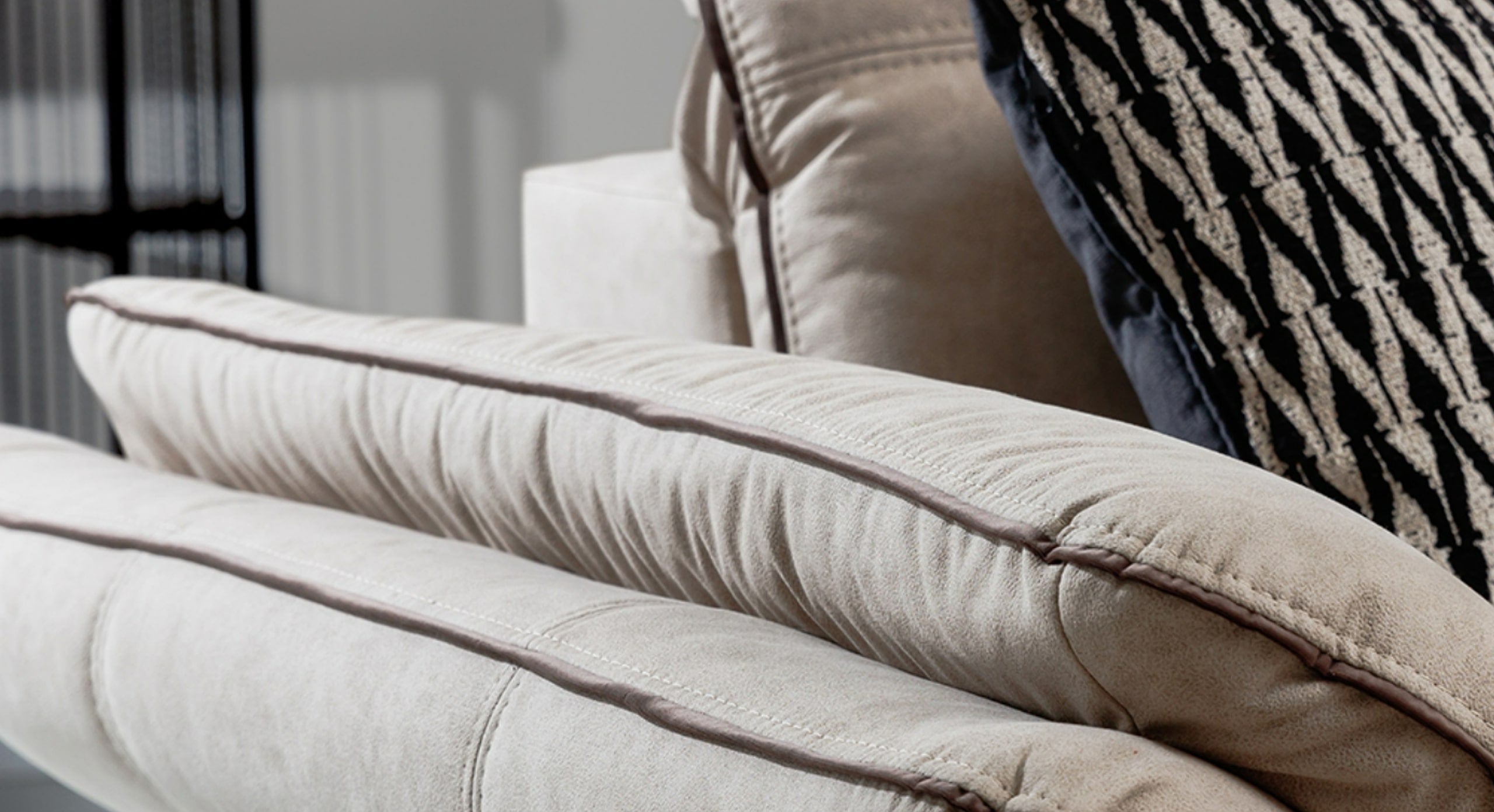 Close-up van de armleuning van de hoekbank Charm met chaise longue, bekleed met beige stof en voorzien van decoratieve biezen voor een verfijnde uitstraling. Op de achtergrond ligt een zwart-wit patroonkussen dat een modern accent toevoegt aan het geheel.