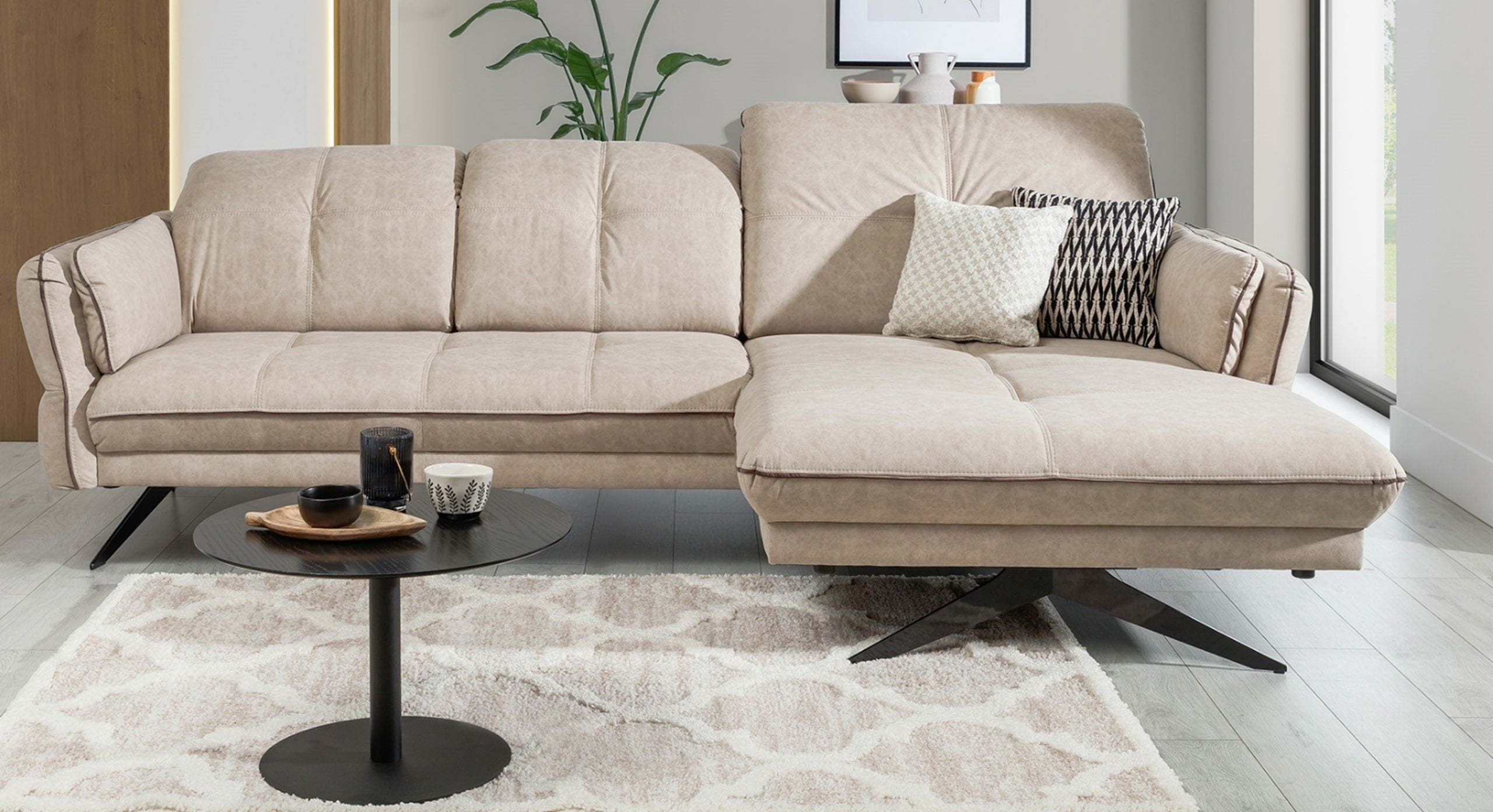 Beige hoekbank Charm met chaise longue rechts, geplaatst in een modern ingerichte woonkamer. De bank heeft zwart metalen poten en sierkussens in patronen voor een stijlvol accent. Voor de bank staat een rond en zwart bijzettafeltje.