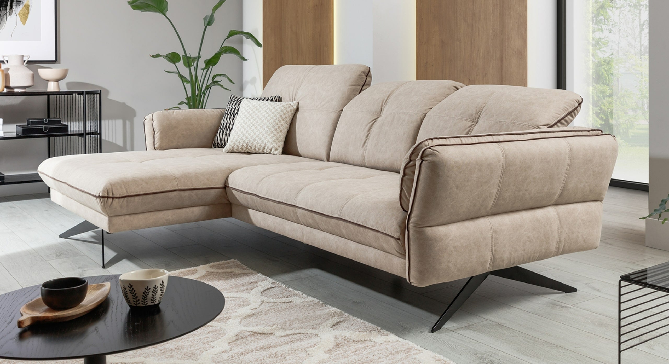 Beige hoekbank Charm met chaise longue links in een modern interieur, voorzien van zwarte metalen poten en gewatteerde, zachte kussens voor extra comfort. De bank is stijlvol gestyled met een contrasterend sierkussen en een bijpassend beige patroonkleed op een lichte vloer.