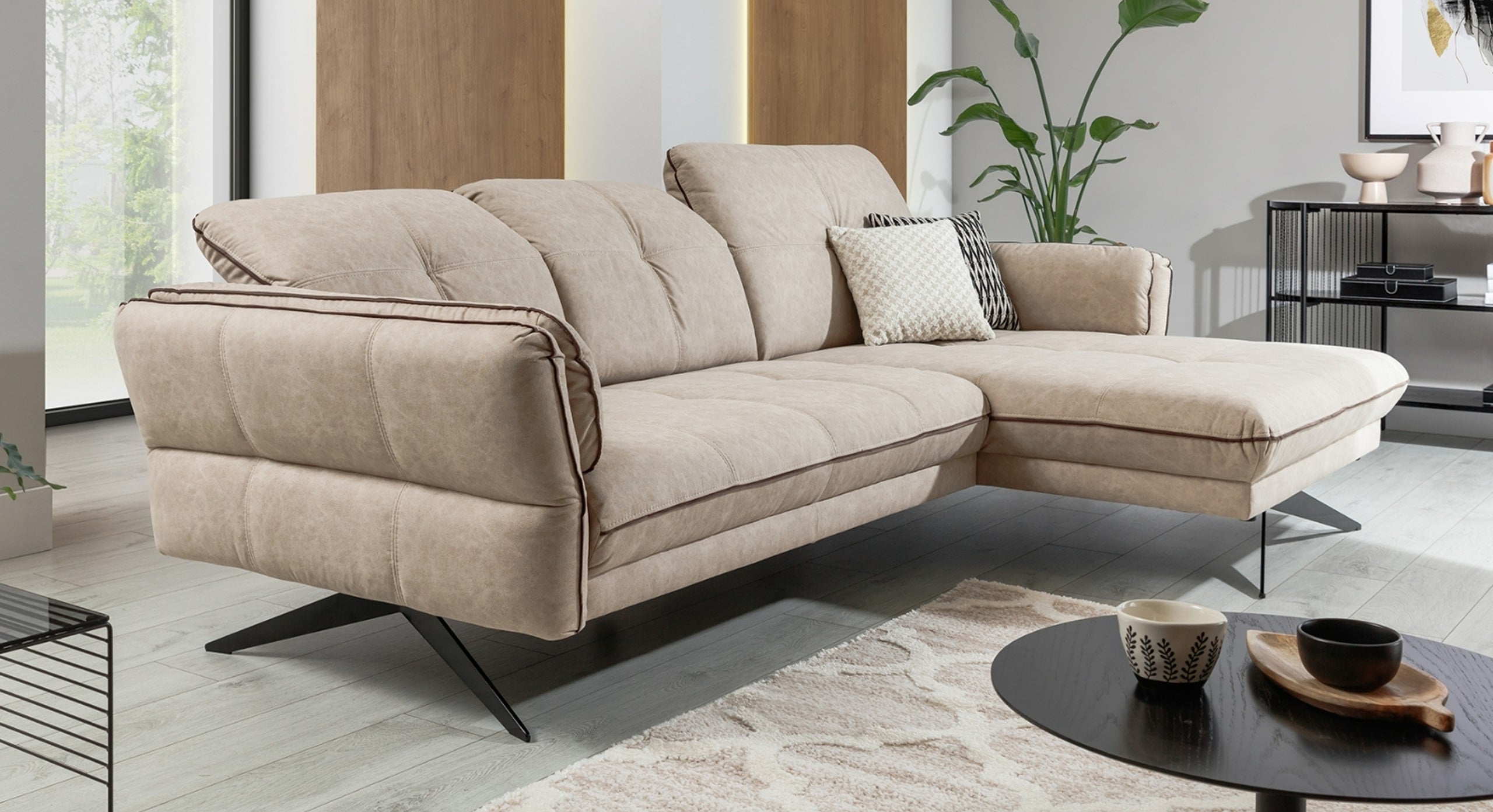 De hoekbank Charm met chaise longue, in een moderne beige stof, toont zijn stijlvolle vormgeving met slanke, zwarte metalen poten en contrasterende biezen langs de zijkanten. In een eigentijds ingerichte kamer biedt de bank een verfijnde uitstraling en comfortabele zitplaatsen.