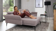 Sfeerbeeld van de Hoekbank Relax met chaise longue links, bekleed in lichtpaarse stof. De moderne woonkamer heeft een warme uitstraling met een grijs vloerkleed en bijzettafel, en de bank biedt comfortabele ruimte voor ontspannen momenten met het gezin.