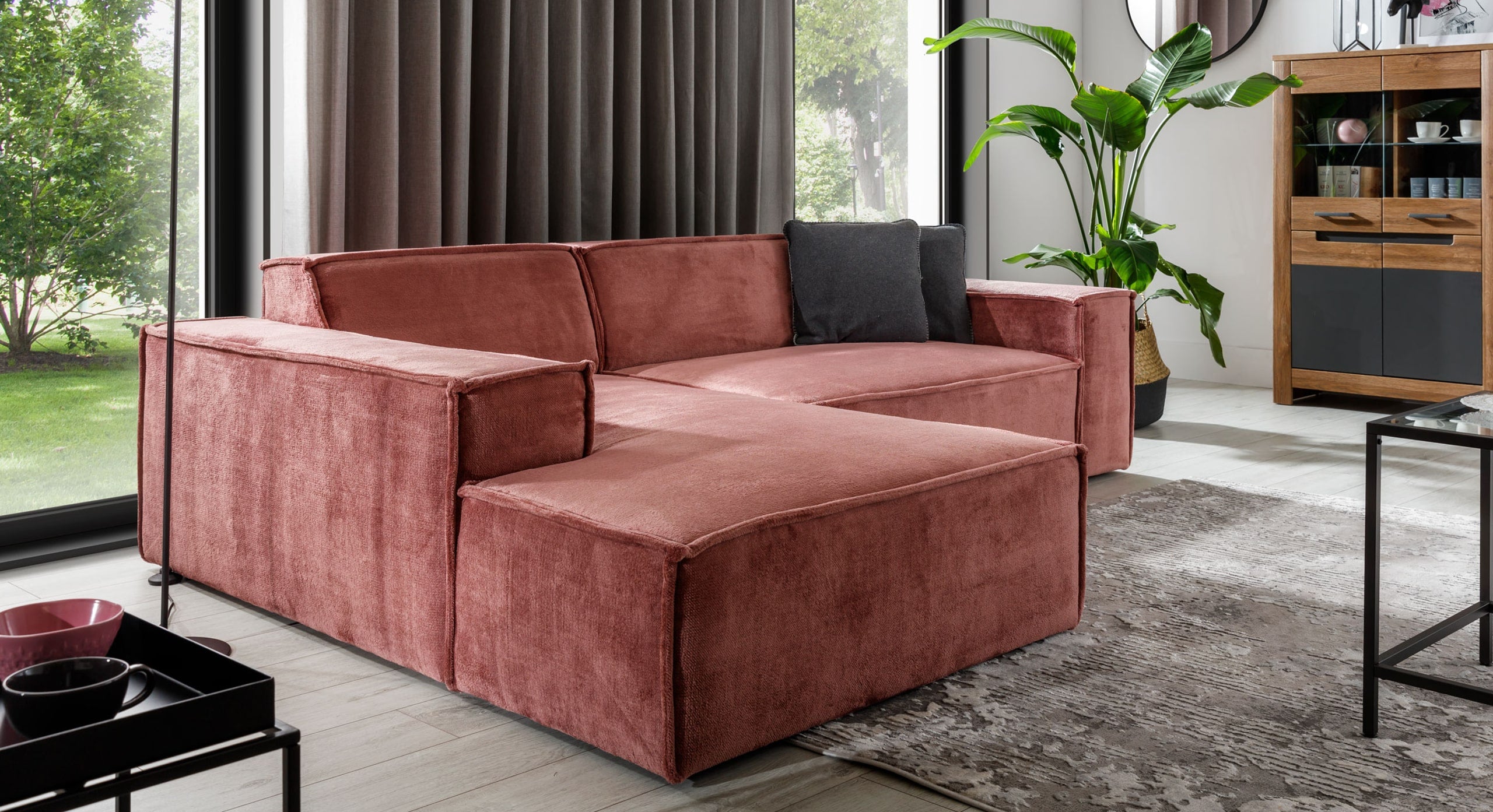 Hoekbank Sense met chaise longue links in een warme roze stof, geplaatst in een moderne woonkamer met veel natuurlijk licht. De strakke lijnen en brede armleuningen van de bank passen perfect bij de neutrale kleuren en groene planten in het interieur, wat zorgt voor een eigentijdse en uitnodigende sfeer.