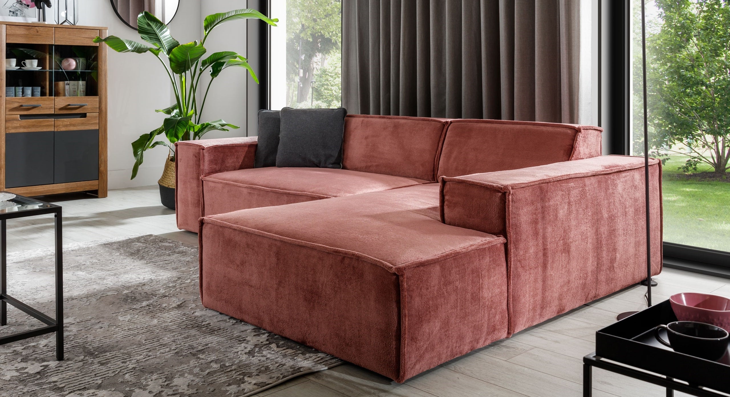 Hoekbank Sense met chaise longue rechts in een warme roze stof, geplaatst in een moderne woonkamer met veel natuurlijk licht. De bank is gecombineerd met een vloerkleed en groene plant, wat de ruimte een eigentijdse en uitnodigende sfeer geeft.