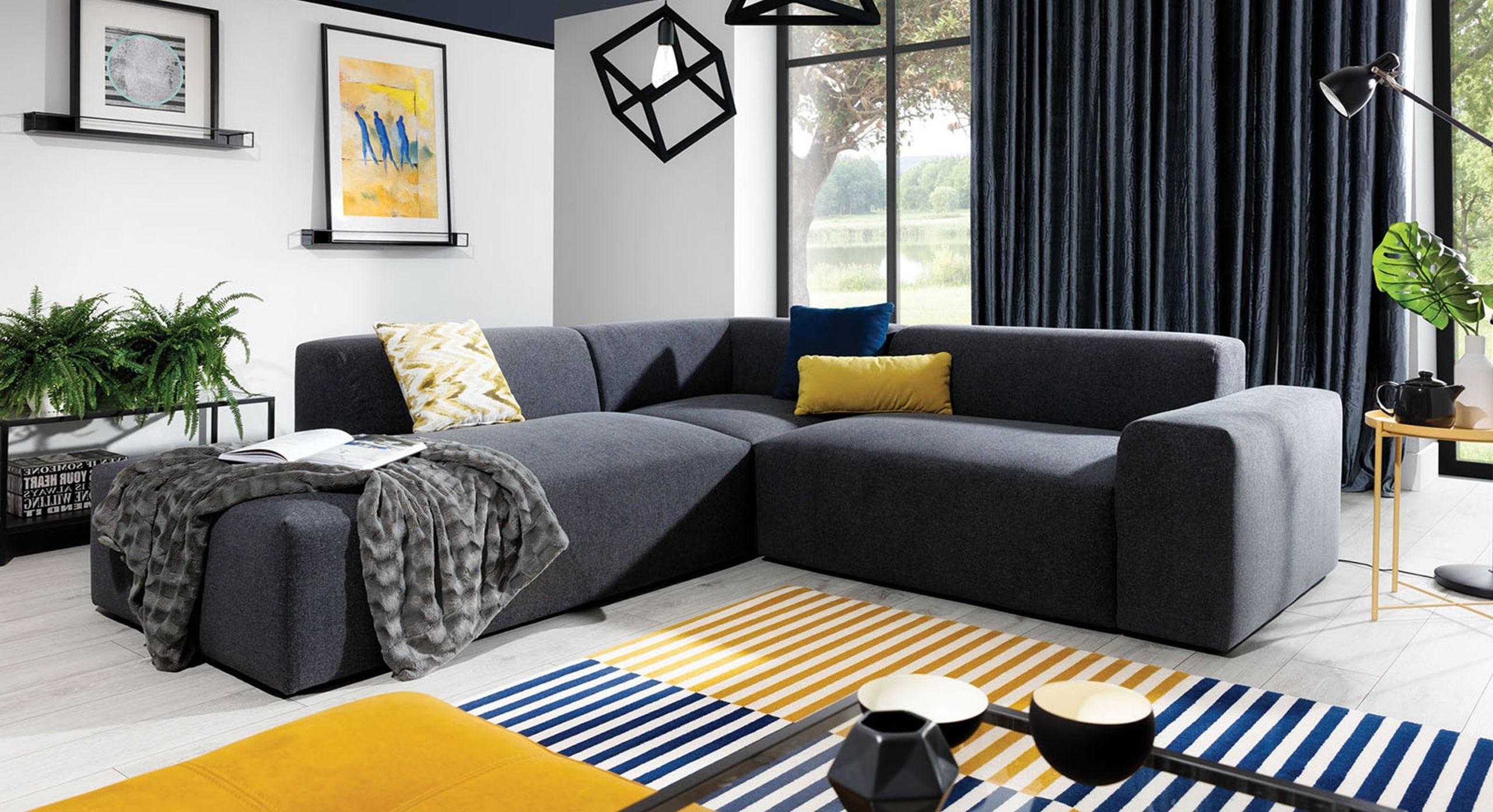 Hoekbank Vibe met hoek en eiland links, afgebeeld in een stijlvolle en moderne woonkamer met een donkere bekleding. De bank is gedecoreerd met kussens in geel en blauw, een plaid, en omgeven door kleurrijke accessoires en geometrische patronen, wat zorgt voor een levendige en eigentijdse sfeer.