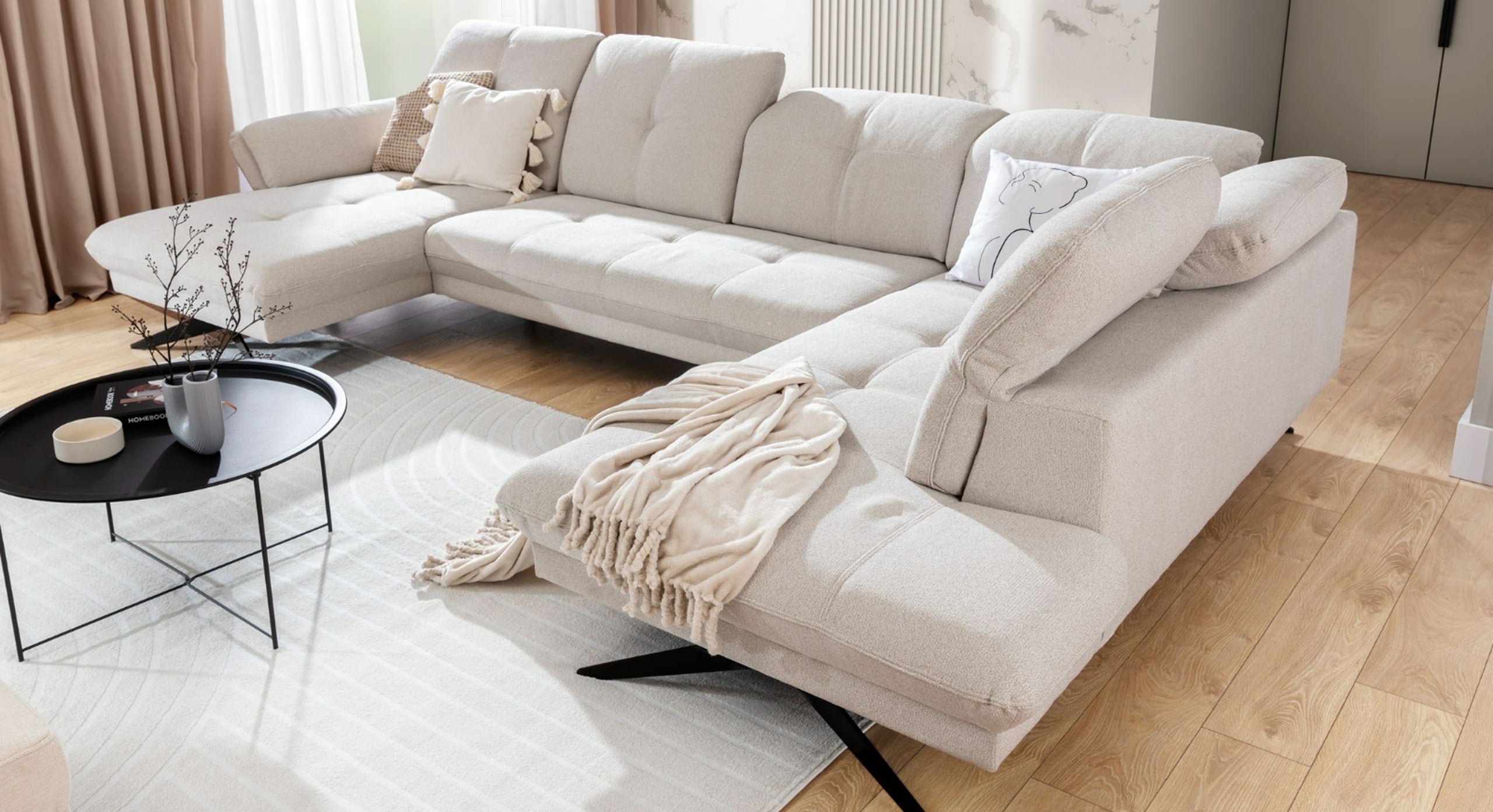 U-bank Charm met chaise longue links, afgebeeld in een lichte en stijlvolle woonkamer. De bank is aangekleed met kussens en een zacht plaid, en wordt vergezeld door een zwarte ronde salontafel met decoratieve elementen, wat een warme en uitnodigende sfeer creëert in het interieur.