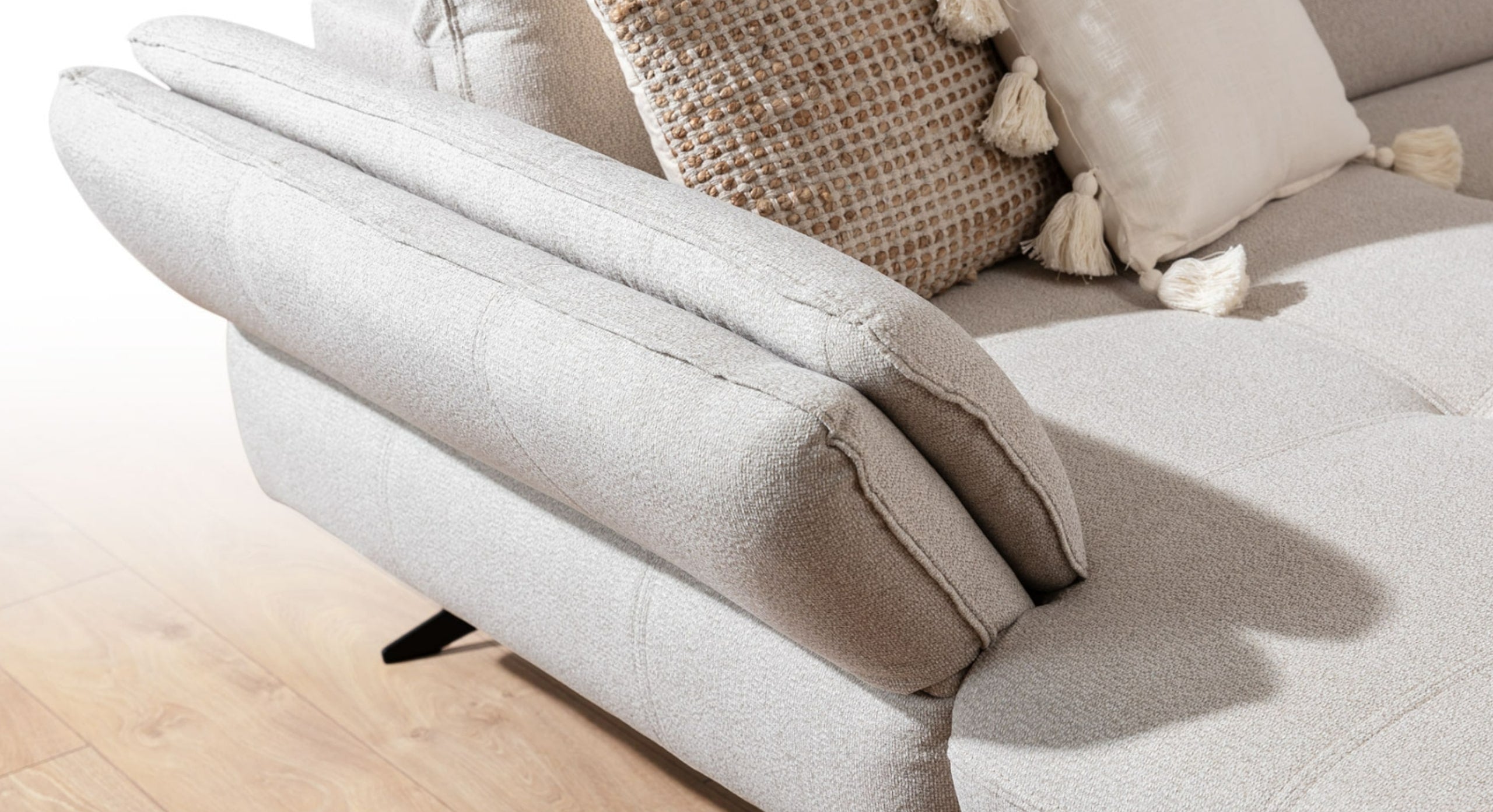 Close-up van de armleuning van de U-bank Charm met chaise longue links, in een lichte stof. De bank is gestyled met een decoratief kussen met kwastjes, wat een extra sfeervol en gezellig accent toevoegt aan het elegante ontwerp.