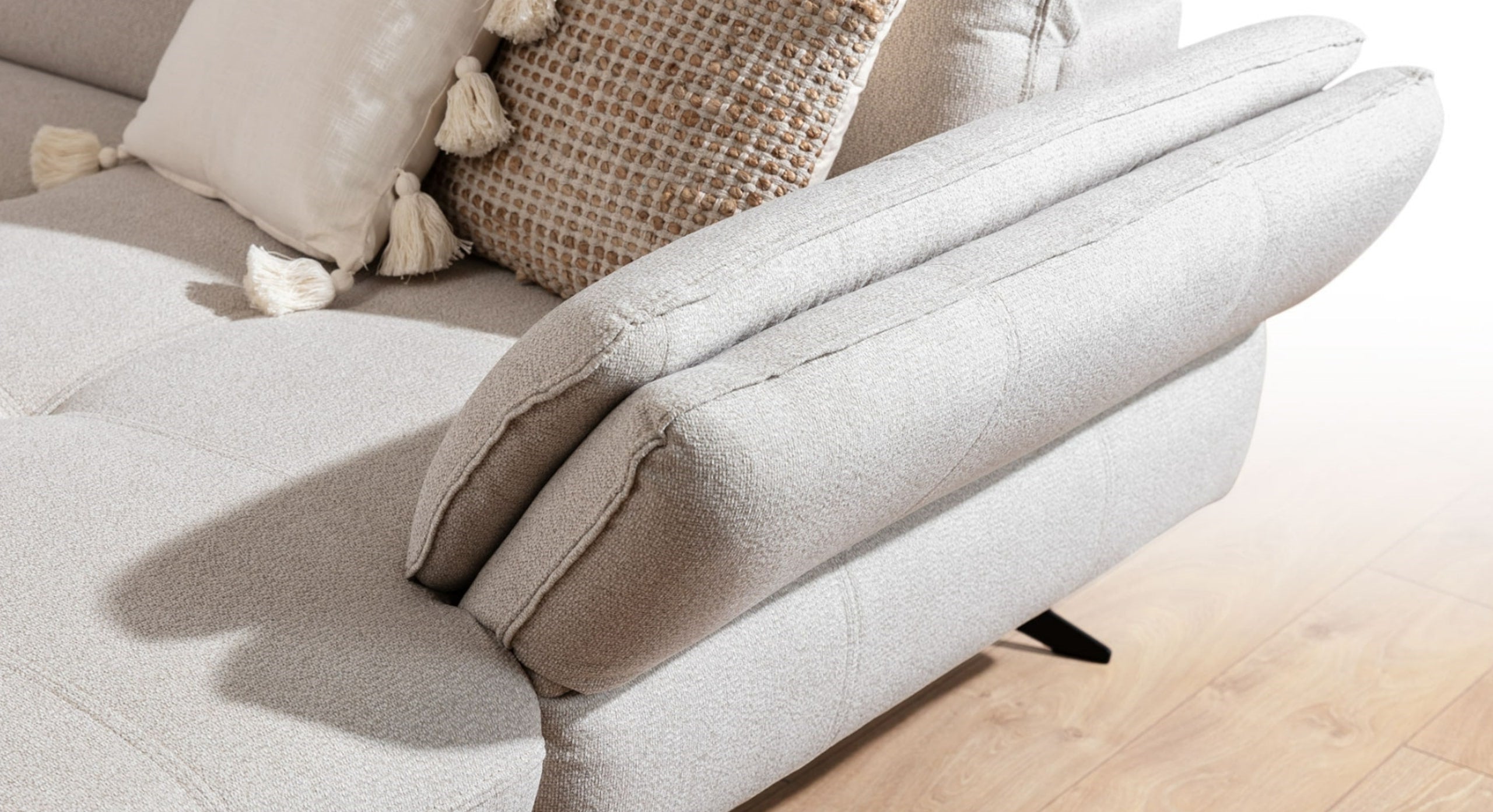 Close-up van de armleuning van de U-bank Charm met chaise longue rechts, in een lichte beige bekleding. De bank is gestyled met een kussen met kwastjes, wat een speels en gezellig accent toevoegt aan het strakke en moderne ontwerp van de bank.