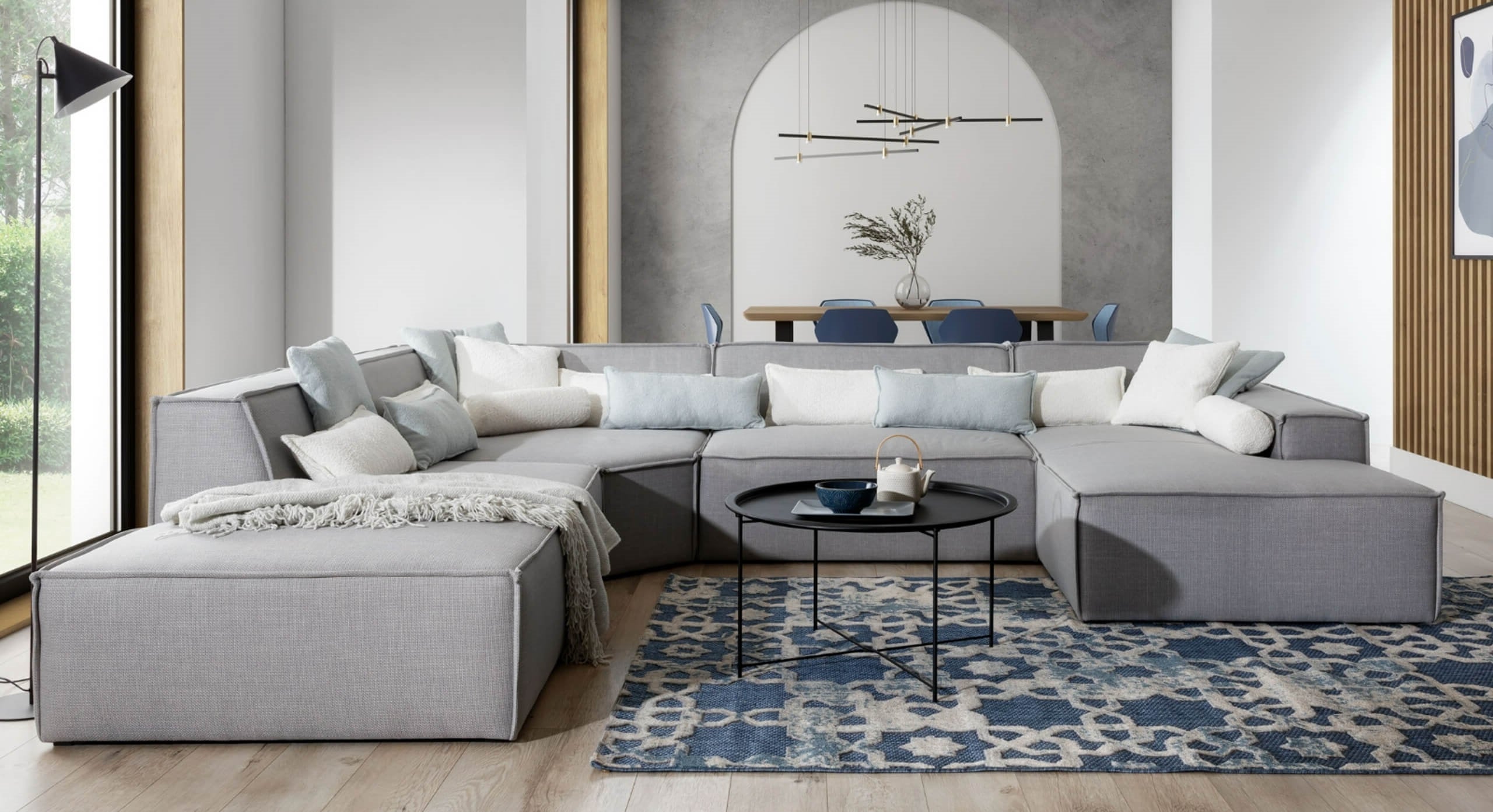 U-bank Sense met chaise longue rechts, geplaatst in een stijlvolle en ruime woonkamer met een blauw-wit patroonvloerkleed. De grijze bank is gedecoreerd met kussens in witte en lichtblauwe tinten en een plaid, en wordt gecombineerd met een zwarte ronde salontafel, wat zorgt voor een elegante en uitnodigende sfeer in het interieur.