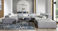 U-bank Sense met chaise longue links, geplaatst in een ruime, moderne woonkamer met een blauw en wit patroonvloerkleed. De bank is gestyled met witte en lichtblauwe kussens en een zwarte ronde salontafel, wat een frisse en uitnodigende uitstraling creëert in een eigentijdse woonomgeving.